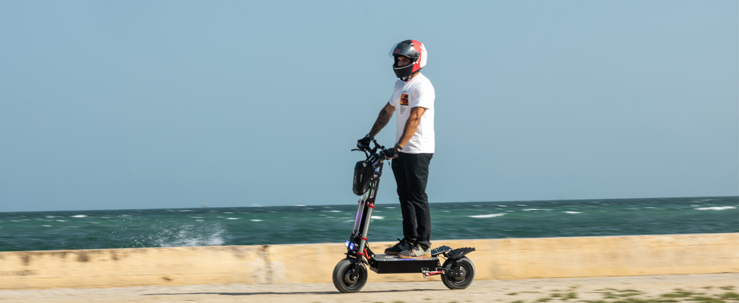 Osoba jeźdząca hulajnogą elektryczną na plaży. Jeździec w białej koszuli i hełmie, piasek i ocean widoczne w tle.